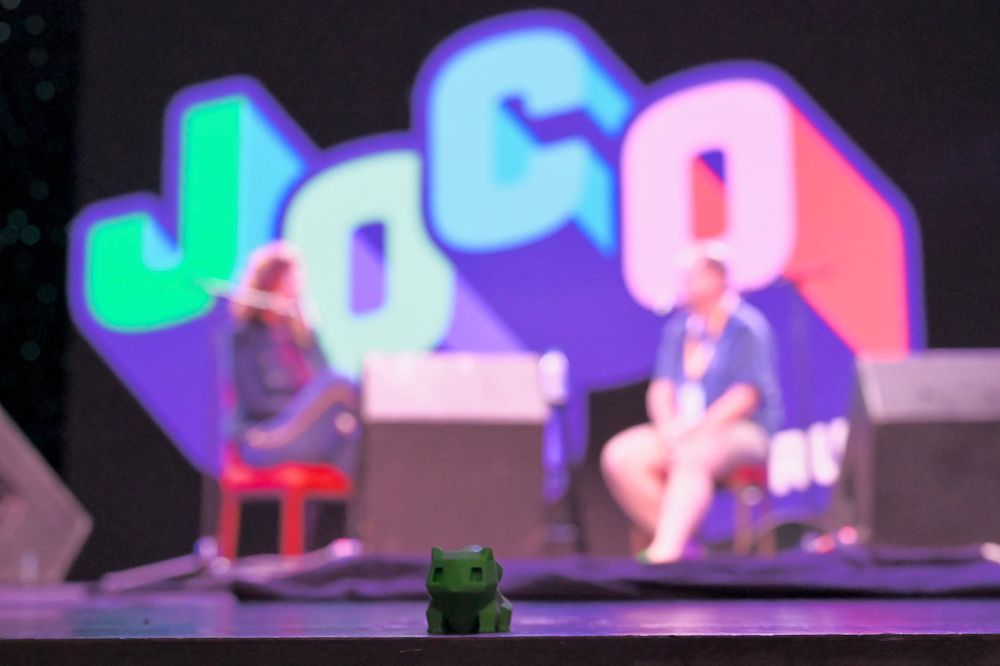 A 3D printed green bulbasaur is in the foreground onstage with speakers and a large JoCo sign blurred in the background