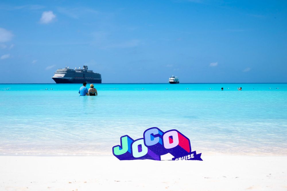 The beach at Half Moon Cay, with white sand in the foreground and blue sea in the back. The JoCo Cruise is pictured in the background, and the official logo (a cut out) is tucked into the sand in the foreground