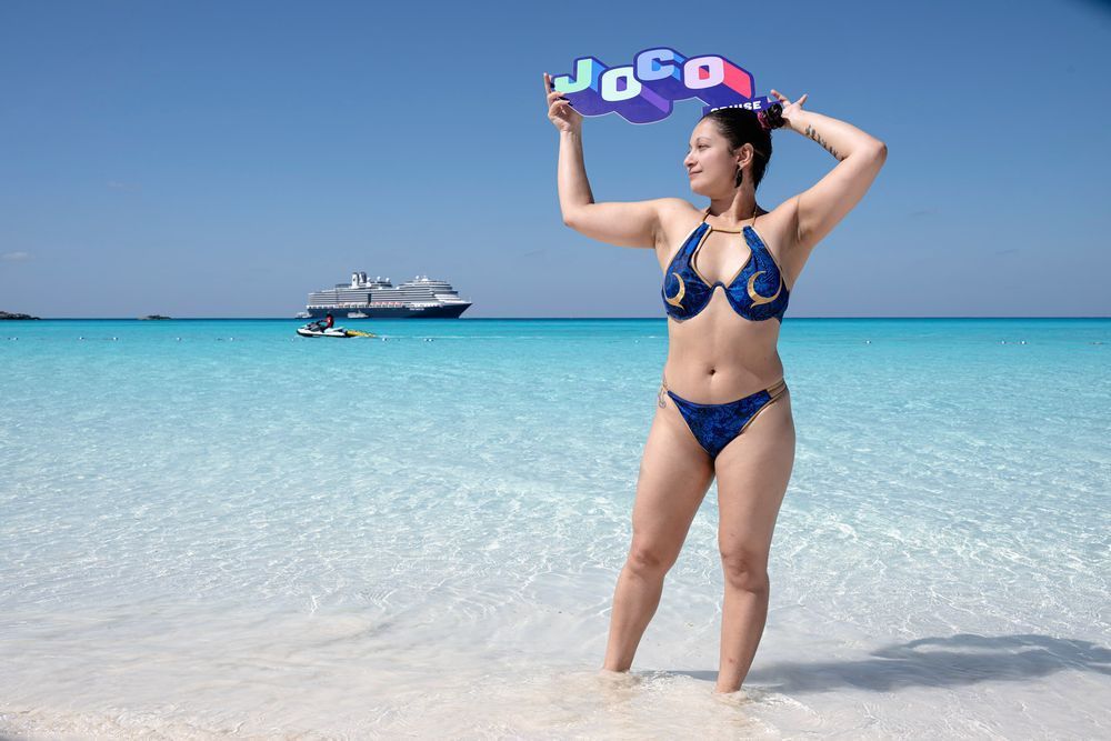 A person wearing a blue Chun Li inspired bathing suit holds a JoCo Cruise sign. They stand in the water of Half Moon Cay, with the cruise ship in the background. The blues of the water and sky are stunning