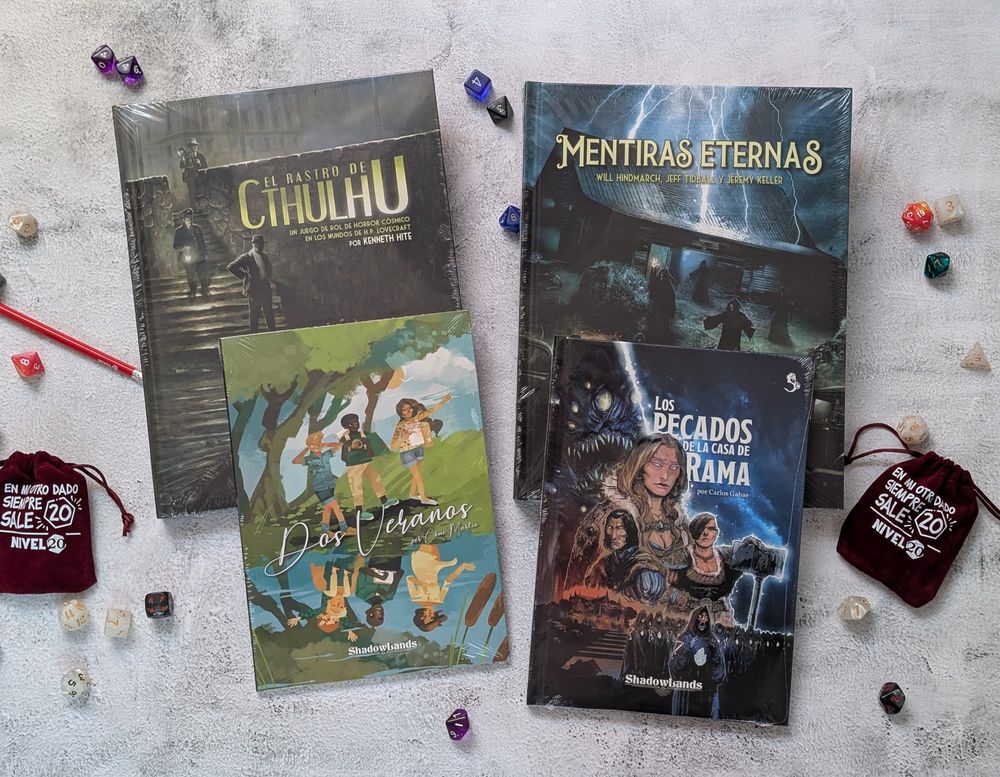Fotografía de los manuales de El Rastro de Cthulhu, Mentiras Eternas, Dos Veranos y el escenario para 5e de Los pecados de la casa de Rama, apoyados sobre una mesa, con dados y bolsas de dados alrededor, sobre un fondo de mármol blanco.
