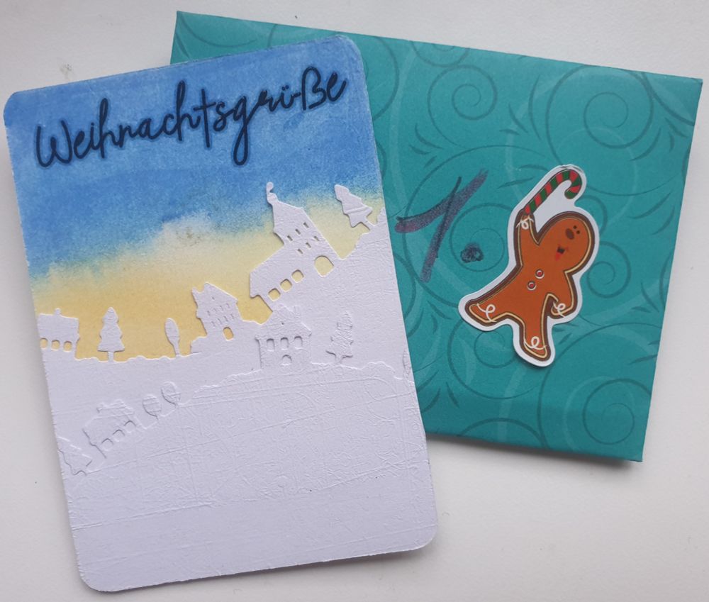 Eine ATC mit zwei gestanzten Haus Silhouetten in weiß auf gelb-blauem Grund. Darüber die Aufschrift "Weihnachtsgrüße"