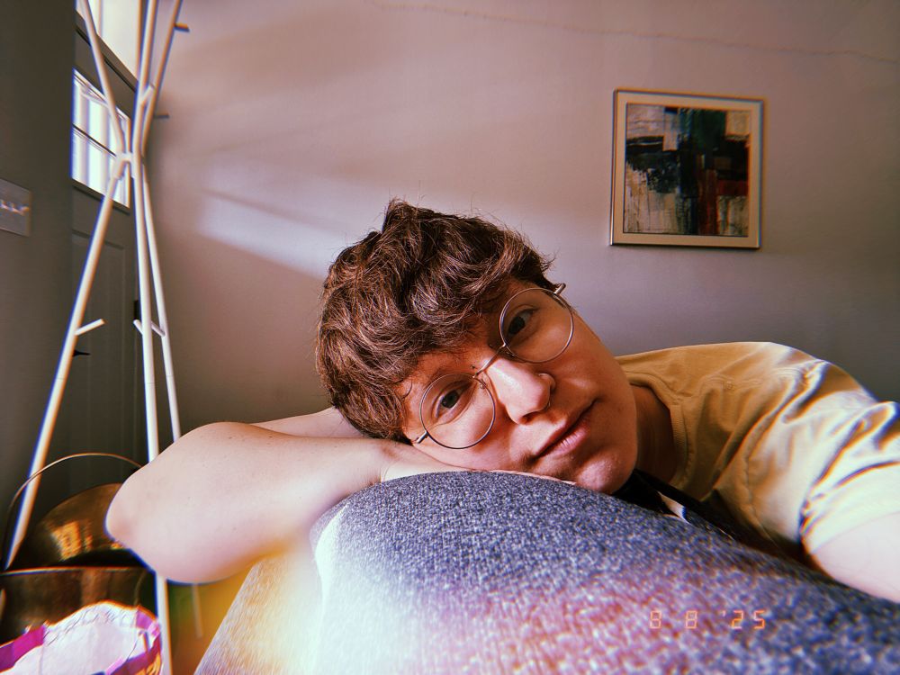 Kari is laying on their hand that’s resting on the back of a couch. They look tired but content. They have messy brown hair that meets their circular framed glasses.