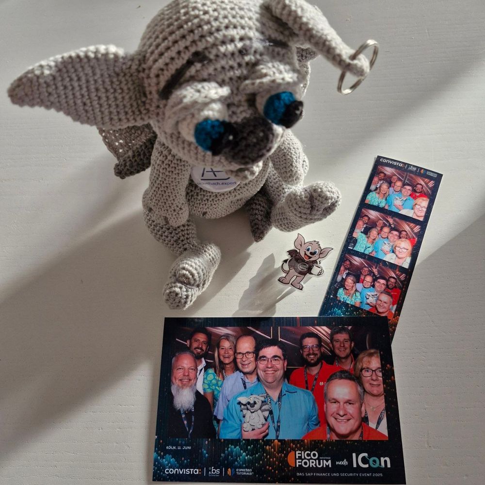 Ein gehäkelter Gargoyle "Gideon" mit großen blauen Augen sitzt auf einem hellen Tisch. Vor ihm liegen ein Fotostreifen und ein größeres Gruppenfoto vom „FICO Forum meets ICON“ Event 2025. Beide zeigen mehrere fröhliche Teilnehmer in bunten Outfits, einer davon hält das Maskottchen in der Hand. Daneben liegt ein kleiner Pin mit dem gleichen Maskottchen in Comicstil. Die Fotos sind mit Logos der Veranstalter „Convista“, „ibs“ und „Espresso Tutorials“ versehen.