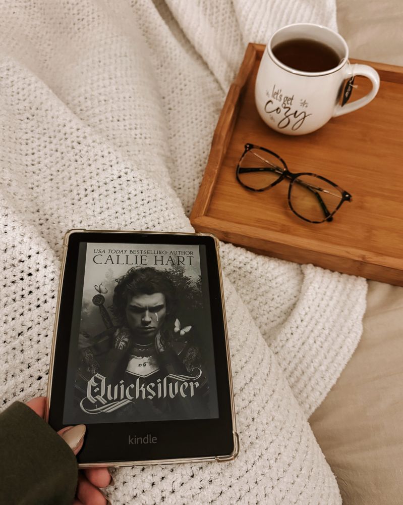 Hand holding kindle with Quicksilver by Callie Hart on it. A cup of tea and a pair of glasses sit on a tray to the side.