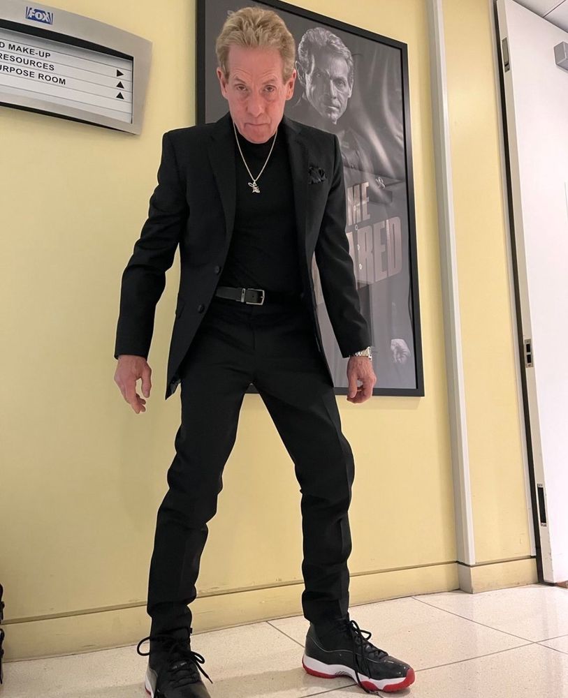 Idiot sports pundit Skip Bayless stanced and fitted in all black with some Jordan sneakers in front of a poster of himself 