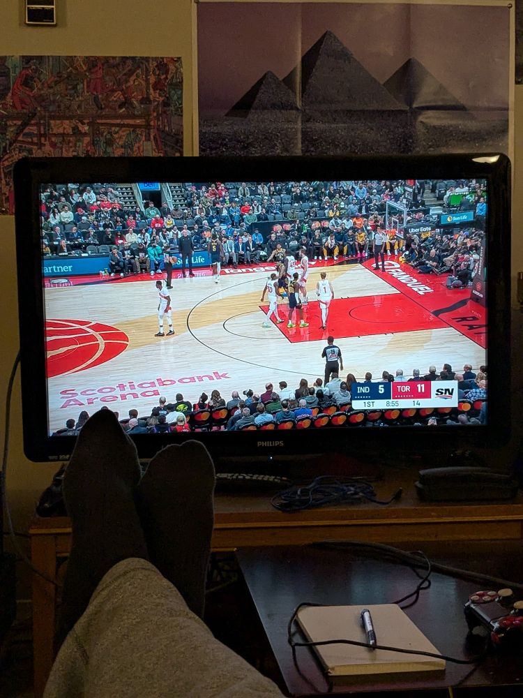 POV of a living room television. A set of reclined human legs, in peak relaxation, occupy the foreground, the Toronto Raptors vs Indana Pacers occupy the background. 