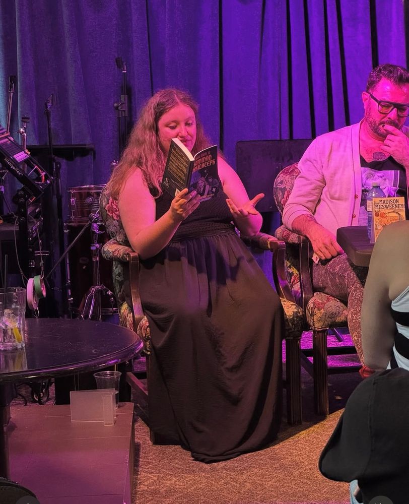 Madison McSweeney in a floor-length black dress reading from Most Likely to Summon Nyhiloteph, seated next to other authors in a room drenched in purple lights 