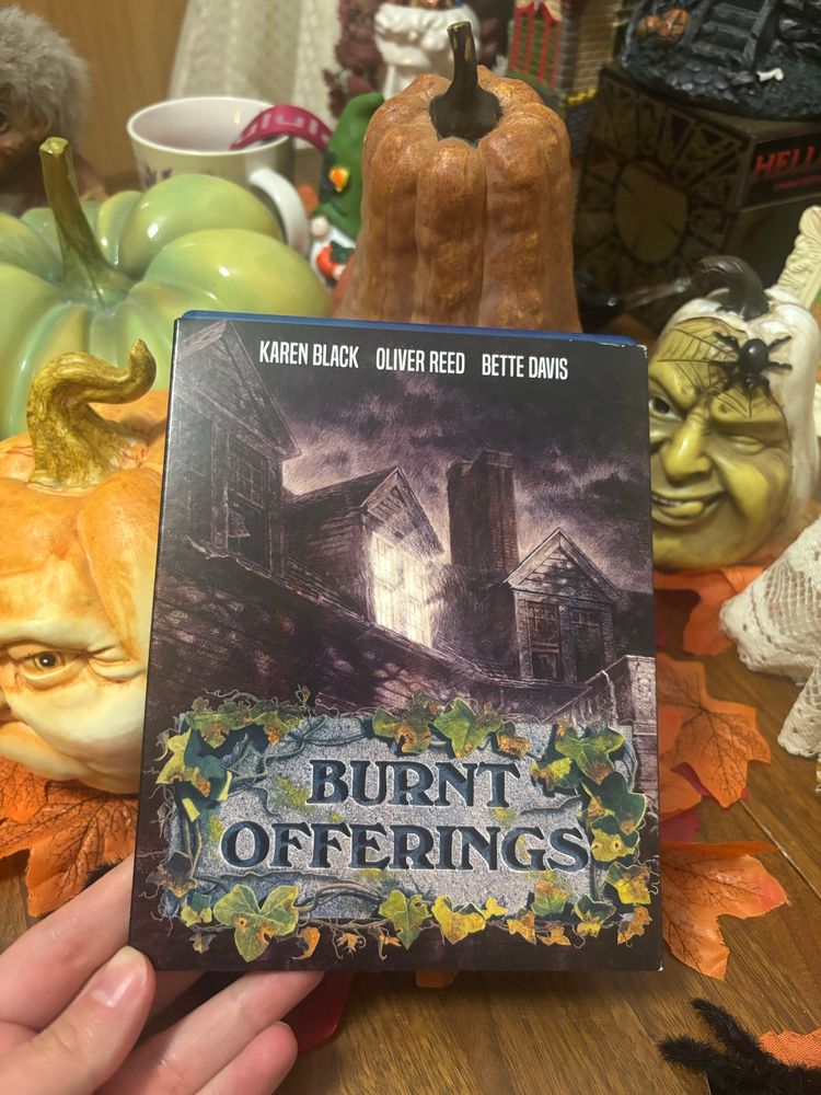DVD cover for Burnt Offerings, depicting the top half of a spooky house with a light in a single upper window, held up against a backdrop of ceramic gourds 