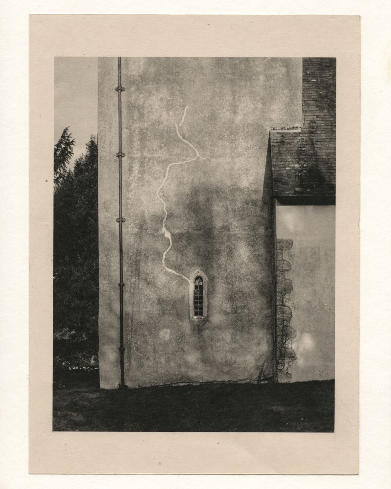 This is a black and white photograph of a 13th century Norman church in Kent, England. The image focuses on a weathered exterior wall with a small, arched window in the center. A prominent crack runs vertically along the wall, creating an organic, meandering line from the top to the bottom.

To the left, there is a vertical drainpipe attached to the wall. The right side of the image includes part of another section of the building with a sloped roof covered in tiles. Some vegetation is visible growing along the edge of the church. The background contains dark foliage, suggesting a rural or secluded setting.

The photograph is printed on warm toned paper, giving it a vintage or archival feel. The overall composition emphasizes the textures of the weathered wall, the crack, and the contrast between architectural elements and nature.