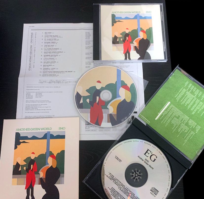 Various copies and editions of Brian Eno's album "Another Green World" on compact discs. There is a disc with part of the album art printed on it in the middle, there is a different disc of the same album in a tray with text on it instead, that one is inside an open CD case with the back of the booklet visible with green on the right. To the left is a "mini LP" edition of the CD which comes in a vinyl-style jacket but smaller. Above it is a foldout with song titles and credits. There is another CD edition in its case on the upper right side. This is all against a black background