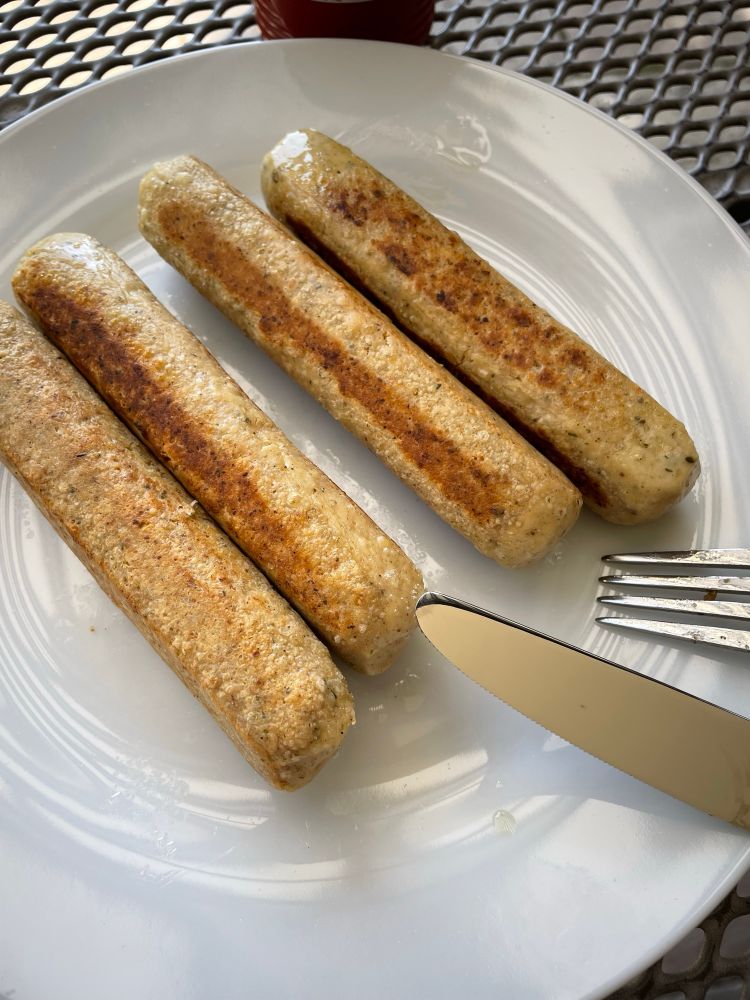Weißer Teller mit 4 Tofu-Würstchen drauf, die gebraten wurden. Rechts  unten ragen Spitze von Gabel und Messer ins Bild. Der Teller steht auf einem Gartentisch aus Metall und hinten/oben ist sehr verschwommen der untere Teil einer Ketchup Flasche im Bild.