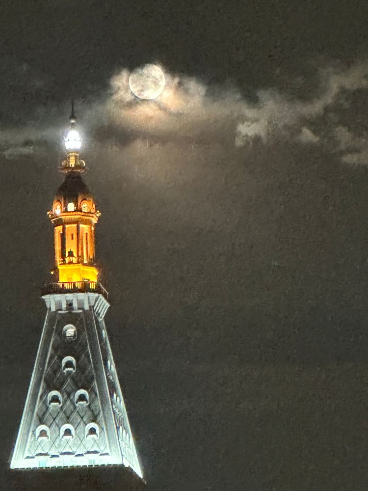 Full moon with some clouds shading it above a tall building lit up in night sky 