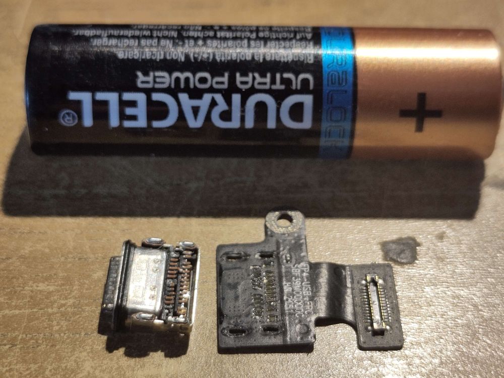 Two tiny electronic parts, with an AA battery for scale which dwarfs them in comparison. The part on the left is a USB-C female port, which the cable plugs into. The part on the right is a flexible circuit board which attaches the USB-C port to the phone via a little push-in connector with many miniscule pins. The right part has some pads and cutouts that were obviously meant to be attached to the left part.