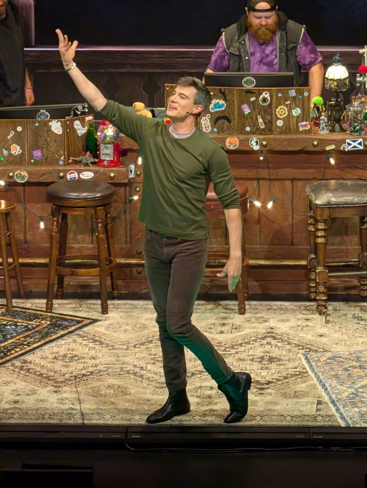 Jacques ze Whipper pushing on stage wearing a green shirt. They are starving in front of a makeshift bar with old wooden stools