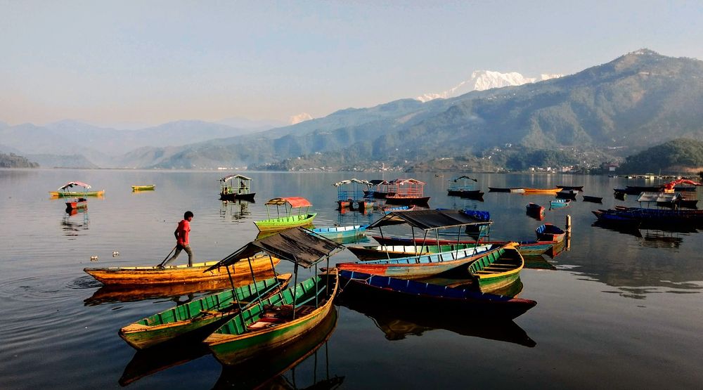 This image shows a serene lake in Pokhara, Nepal, with colorful wooden boats floating on still water. A person walks on one yellow boat, adding life to the scene. In the background, misty green hills and snow-capped mountains rise under a soft, pale sky, reflecting calm beauty.