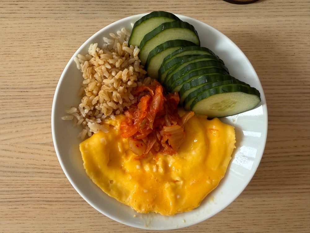 A white bowl containing a yellow scrambled eggs topped with kimchi in the middle, brown rice, and sliced cucumber arranged on a wooden table.​​​​​​​​​​​​​​​​