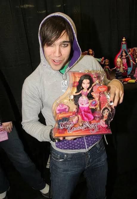 photo of Pete Wentz (fall out boy), he's wearing a gray hoodie with detail in purple and blue jeans. he's holding a set of Bratz dolls.
