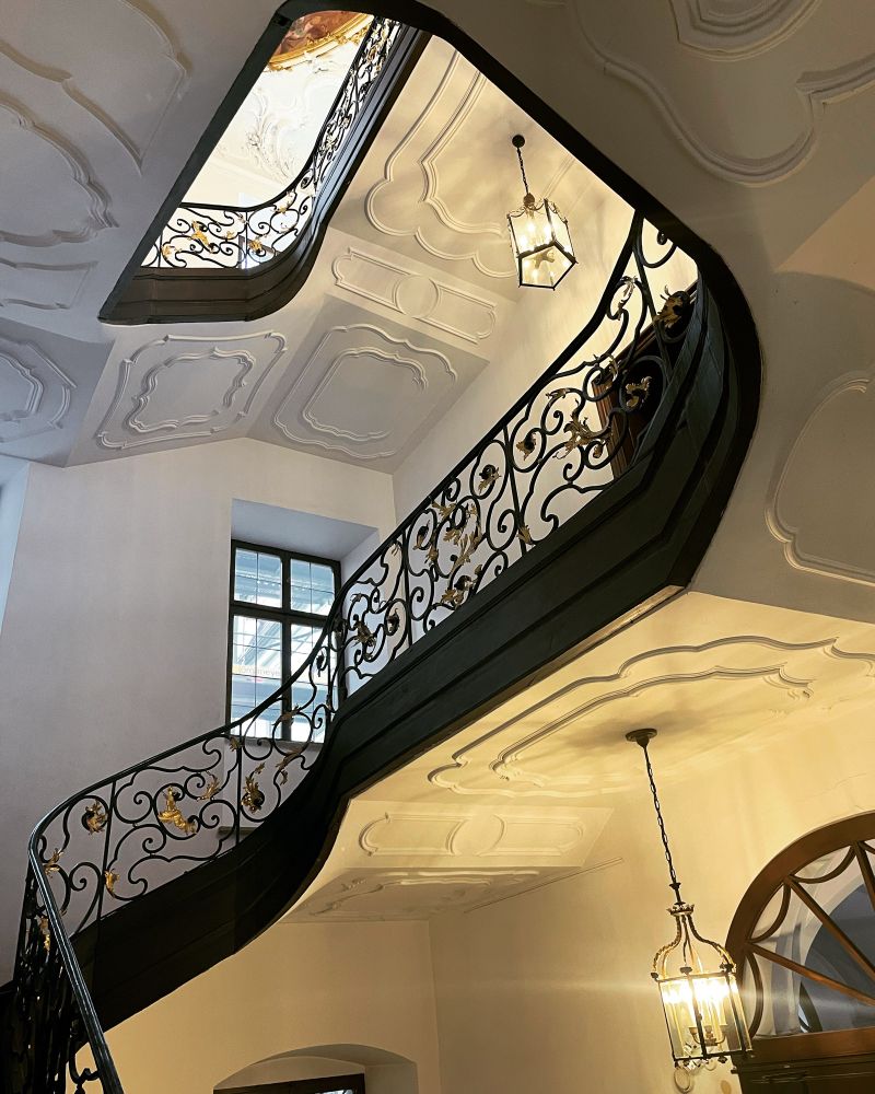 Ein Treppenhaus mit einem Geländer in Schwarz und Gold, mit geschwungenen Ornamenten. 