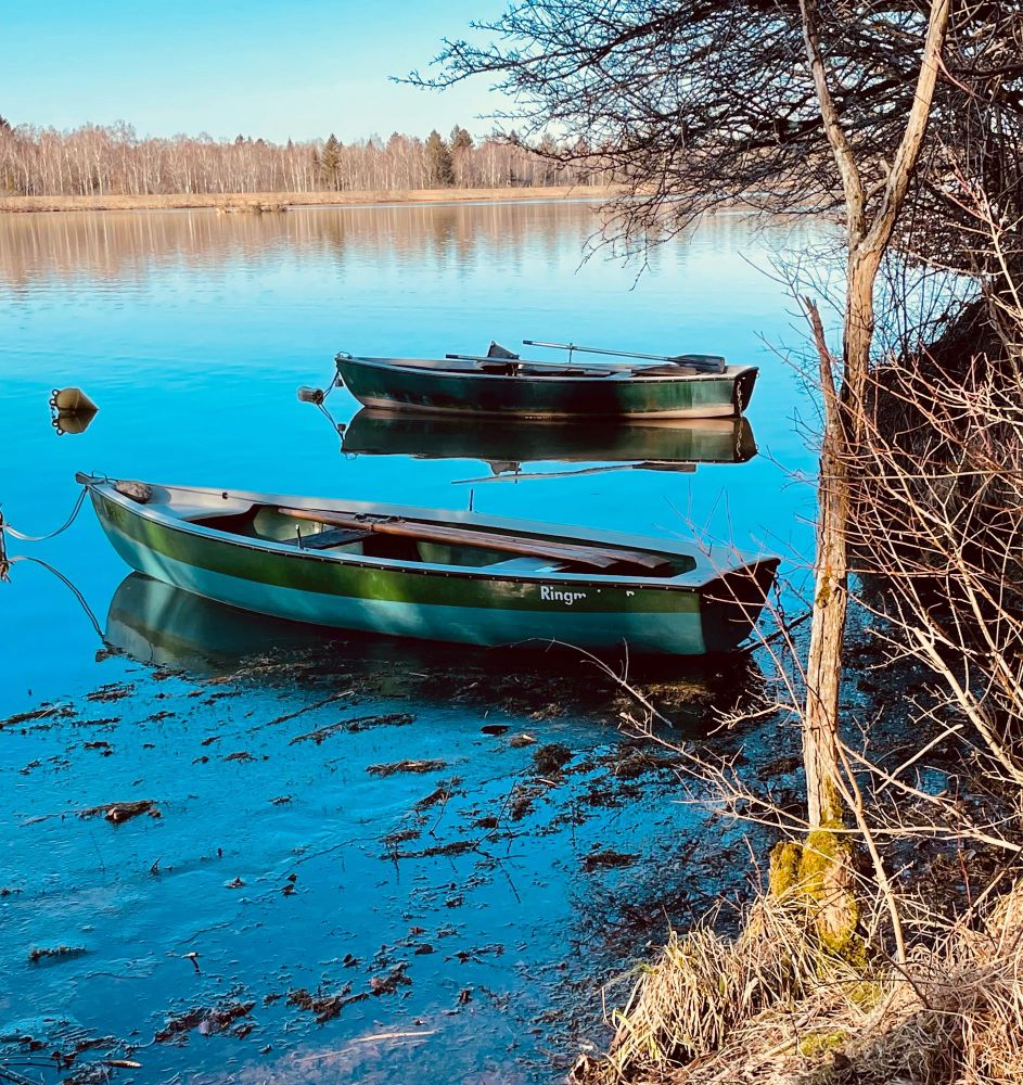 Zwei Boote in Grün auf türkisem Wasser, man sieht vertrocknete Uferböschung am rechten Rand. Im Hintergrund liegt ein Wald.