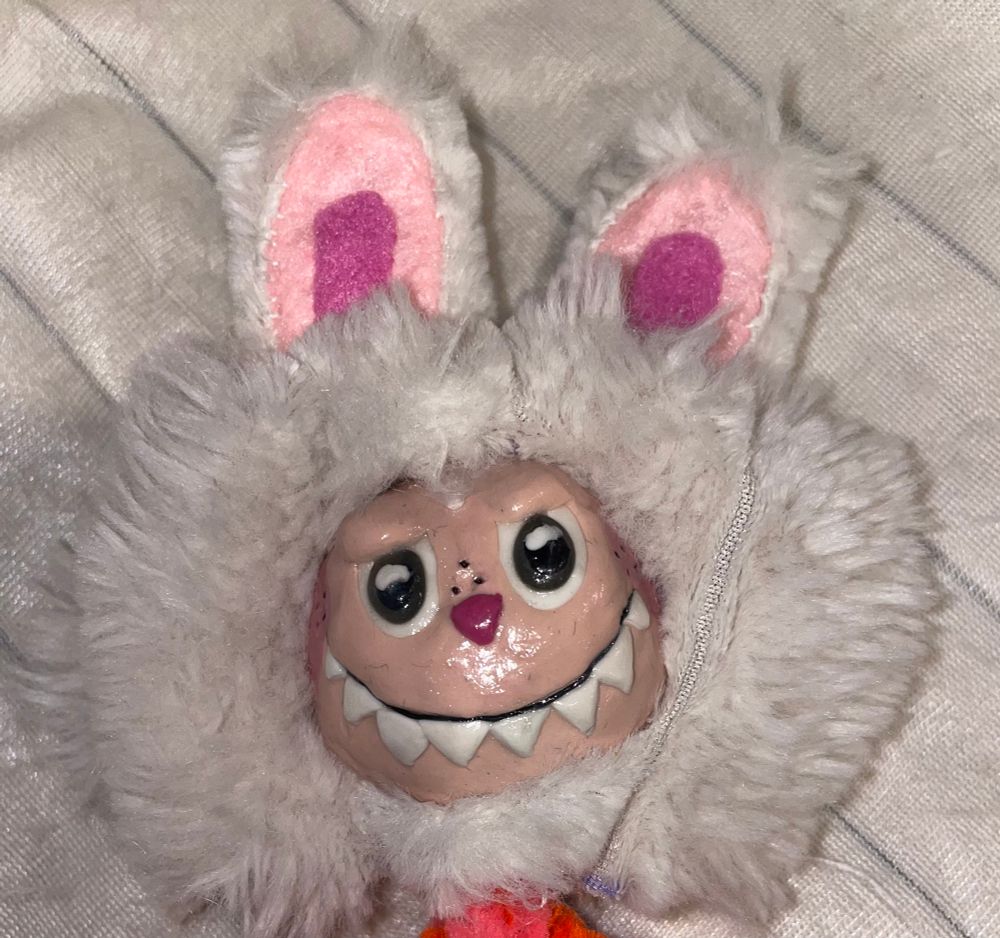 My failed attempt to make a labubu knock-off doll. Picture is just of the head. Creepy face, faux fur, weird bunny ears. On a cream background.