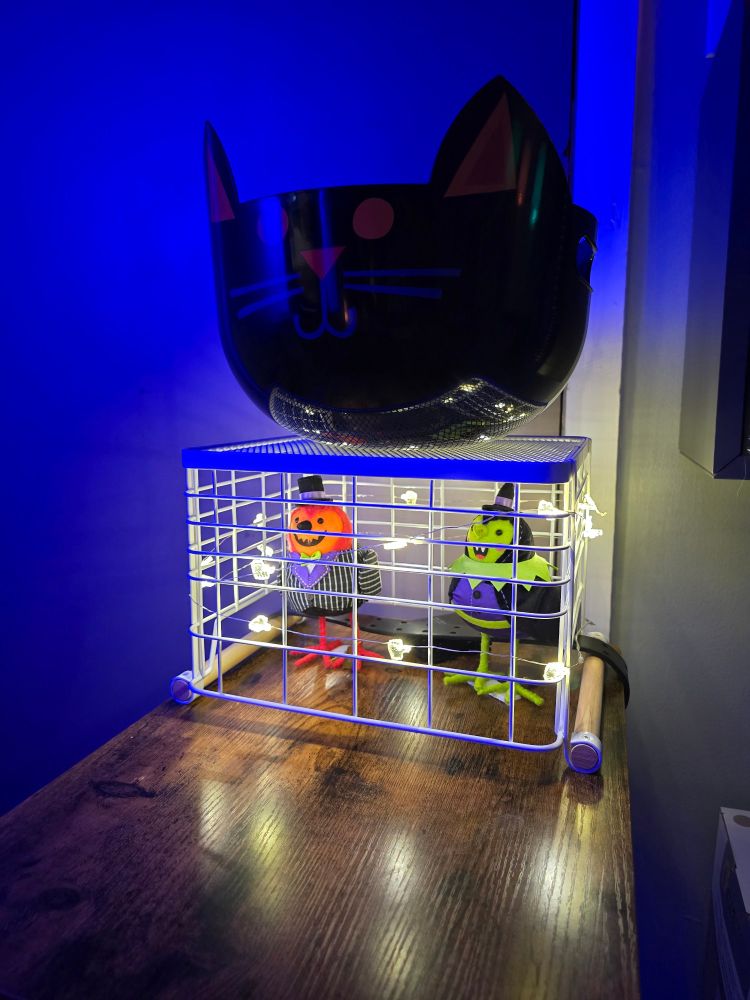 A cat candy bowl sitting on top of a basket wrapped in twinkle lights that is serving as a cage for two decorative Halloween birds. 
