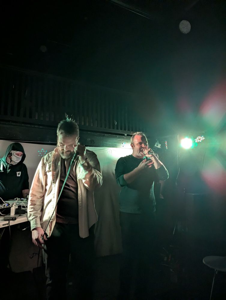 Sam and Joel Herring performing. Sam is on right, rhyming, Joel is gripping the mic and looking down. Deejay Icky Reels is in the background on the boards. 