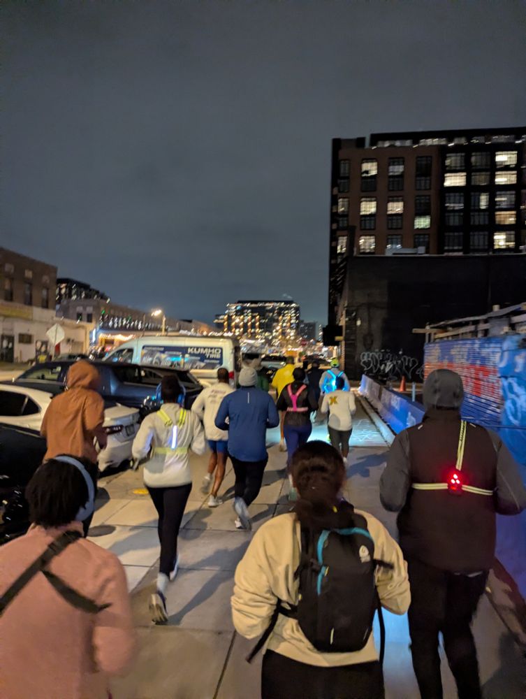 A group of people running down a sidewalk. There are cars parked to the left, a building in the background, and it is night. 