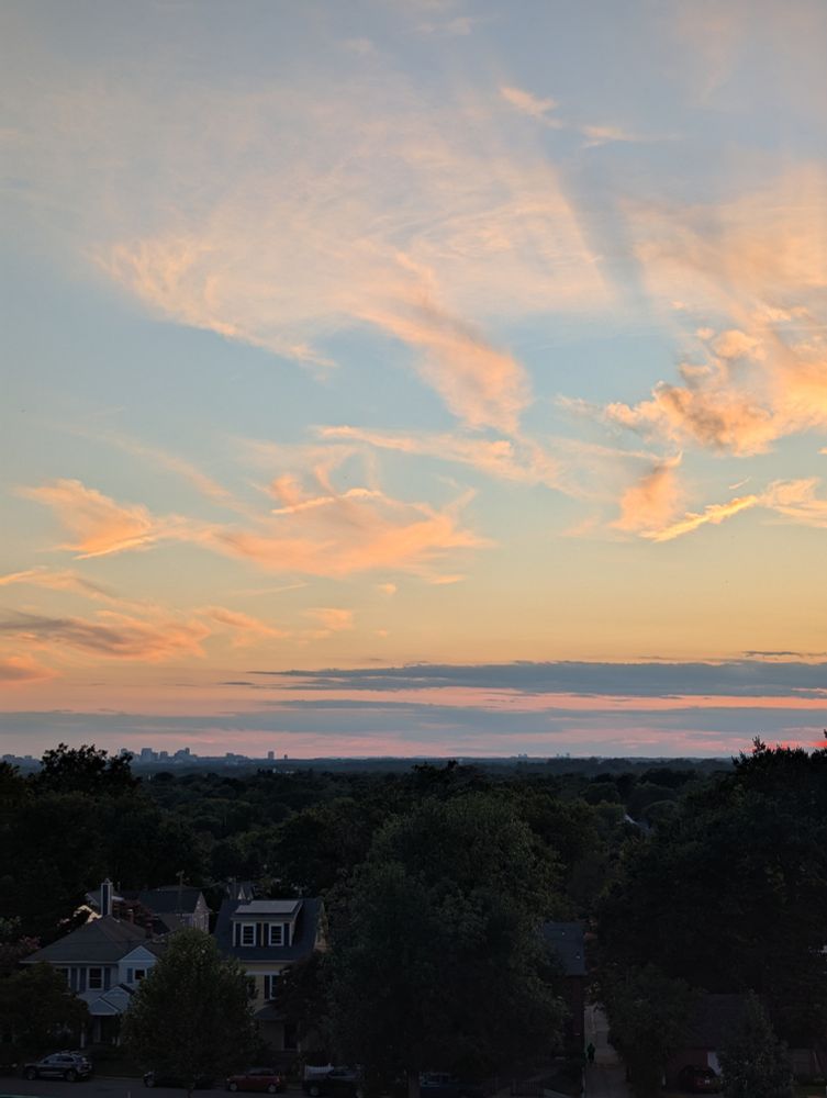 An orange and blue sky with clouds, taken from a hill in NW DC. The sun is red, there are trees and houses in the mid ground. 