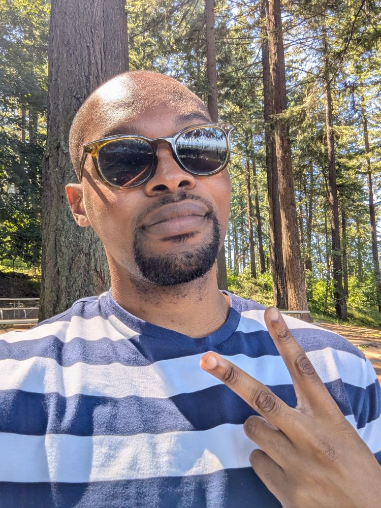 Jazz, a Black man, giving the peace sign while standing under trees. He is wearing sunglasses and a striped blue shirt. 