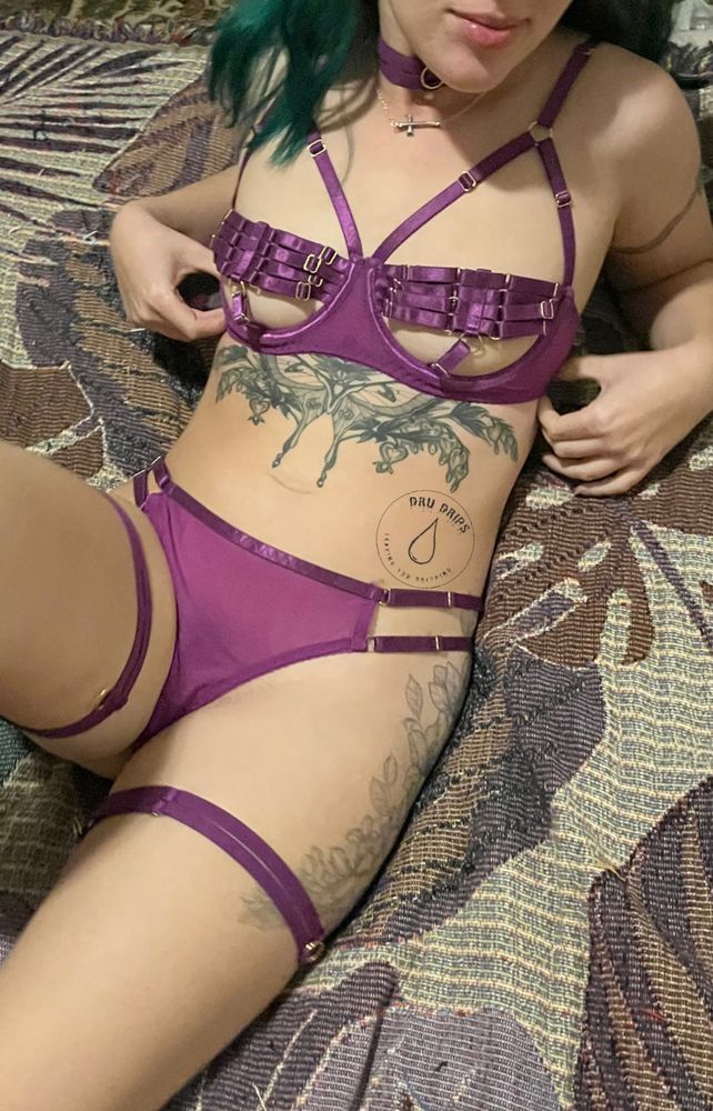 An NSFW queer femme shows off her tattoos and small tits in strappy lingerie with a sweet smile thinking of all the gooners who will fantasize about her