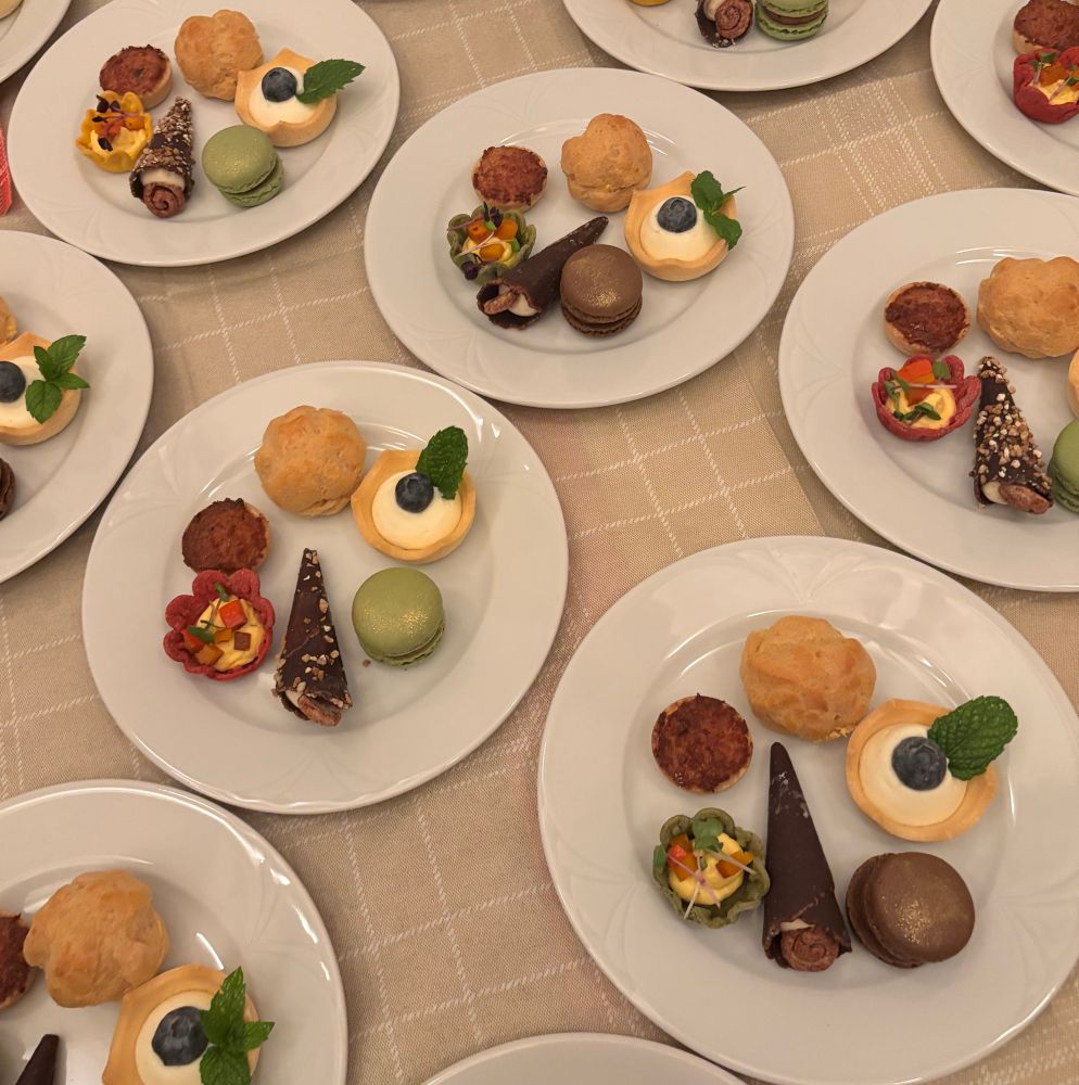 Some plates on a table. Each plate has six small treats on it: a flower shaped tartelette with a squash cream, tiny pumpkin cubes and cress, a small tartelette with a salsiccia filling, a profiterole, a tartelette with a passionfruit cream, blueberry and mint leaf, a macaron with gold dust, a chocolate cone with a caramel filling. 