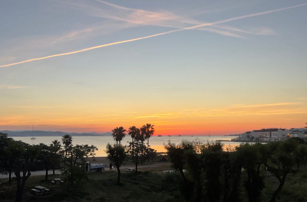 Line of red light burgeoning along the horizon, light reflecting in the still of the bay, pink-tinged contrails crossing the blue sky above.