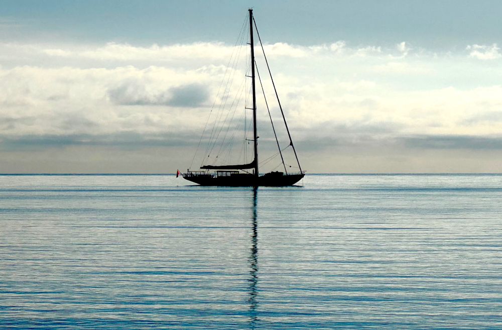 A lone sailing boat at anchor in an empty sea, it’s mast reflecting sharply against the calm surface of the water.