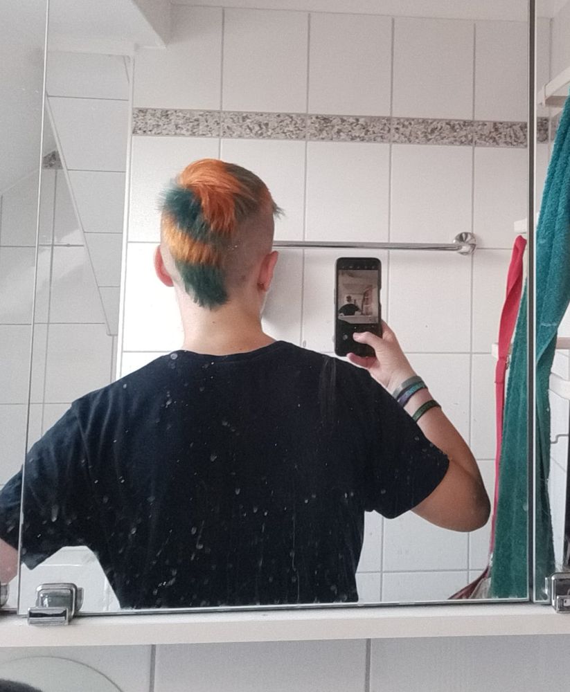 Selfie von hinten in einem weiß gefliesten Bad. Die Person trägt ein schwarzes Shirt und hat einen orange-petrol gefärbten Mohawk-Haarschnitt. (breiter und kürzer als ein Irokesenschnitt)