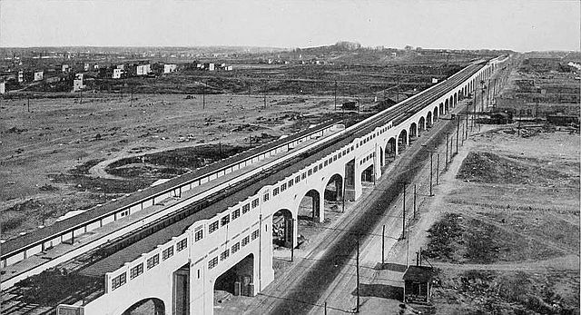 a black and white photo of the 33rd Street–Rawson Street station on the NYC subway in 1920. an elevated track on an arched stone aqueduct stretches into the distance, surrounded by miles of mostly empty dirt 