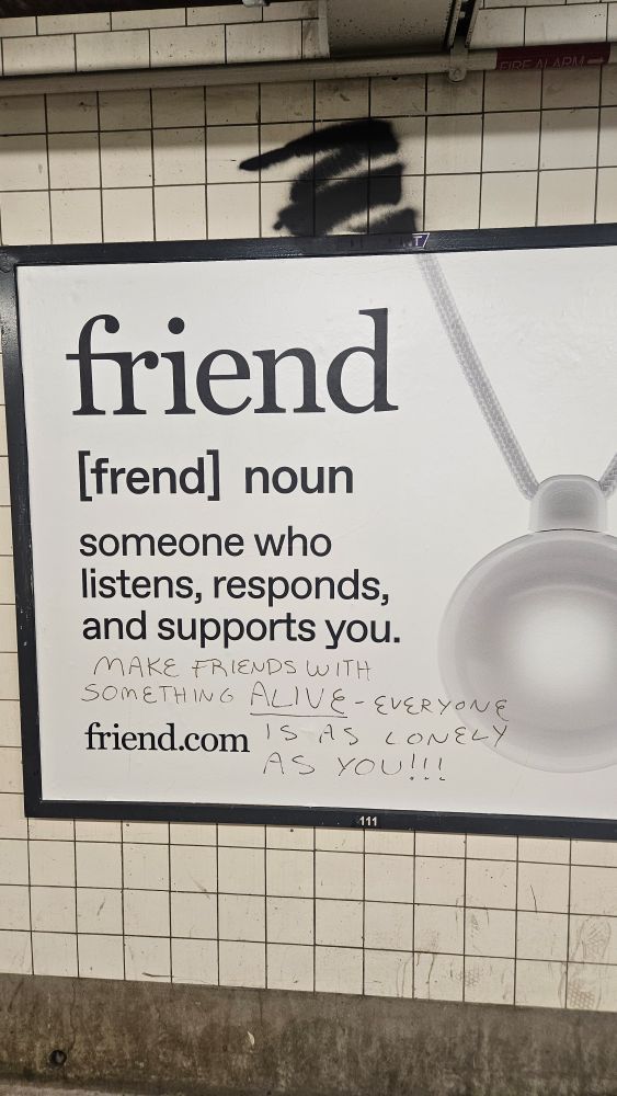 Advertising poster on the white tile wall of a subway station tunnel. The poster features black text on a white background, with the text reading: 
"friend
[frend] noun
someone who listens, tesponds, and supports you.
friend.com"

The right side of the poster also shows an image of a white medallion.  

On the bottom of the poster is handwritten graffiti reading "make friends with something ALIVE - everyone is as lonely as you!!!"