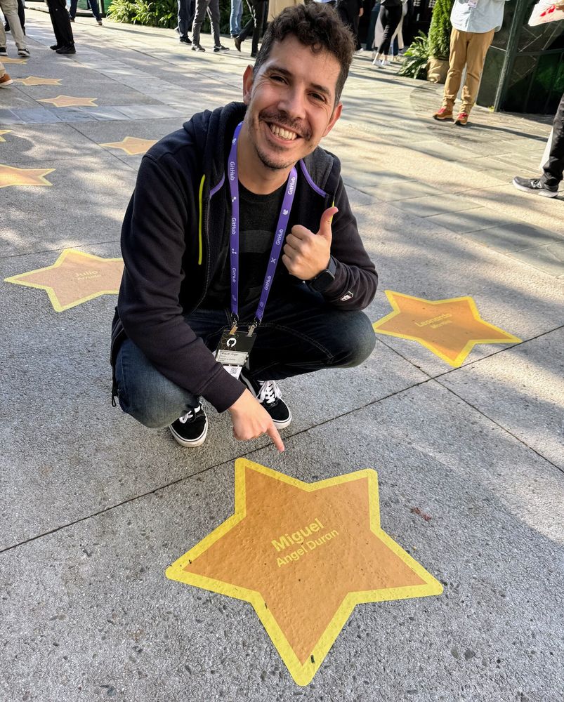 midudev con acreditación de GitHub colgada al cuello se agacha en la calle, hace un gesto de pulgar arriba y señala una estrella amarilla en el suelo que lleva escrito su nombre, como si fuera parte de un paseo de la fama versión programadores.