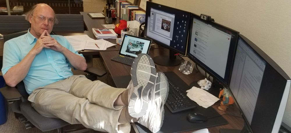 El creador de C++, Bjarne Stroustrup, con camisa turquesa reclinado en su silla de oficina con los pies apoyados en el escritorio, mostrando las suelas de sus zapatillas mientras observa pensativo varios monitores, una tablet y un caos organizado de papeles, libros y tazas – como un ejecutivo estratega en modo descanso.