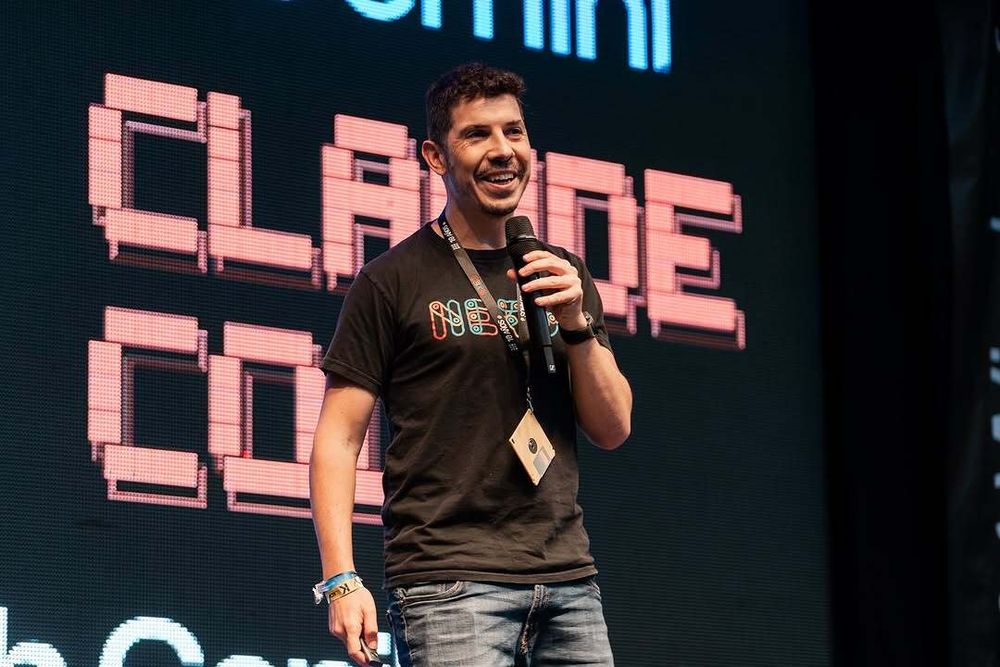 midudev en el escenario de Nerdearla, sosteniendo un micrófono y sonriendo, con una pantalla de fondo con letras pixeladas.