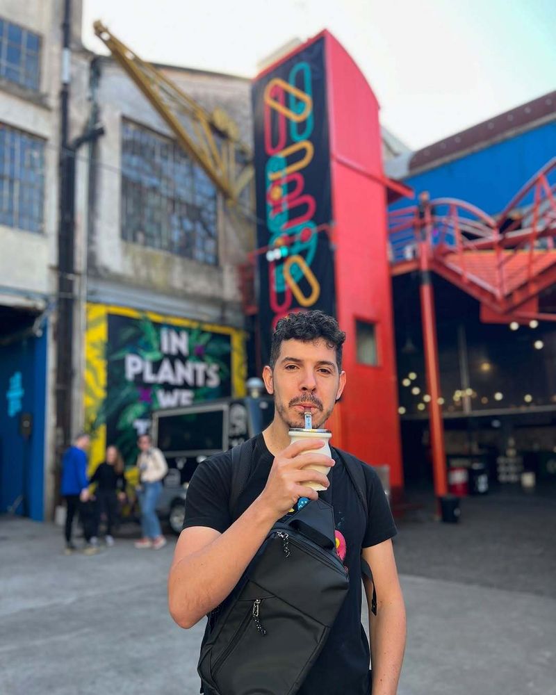 Midudev de pie en el KONEX de Buenos Aires, tomando un mate con bombilla. De fondo se ve el cartel colorido con la palabra “NERDEARLA” y una estructura roja con escaleras.