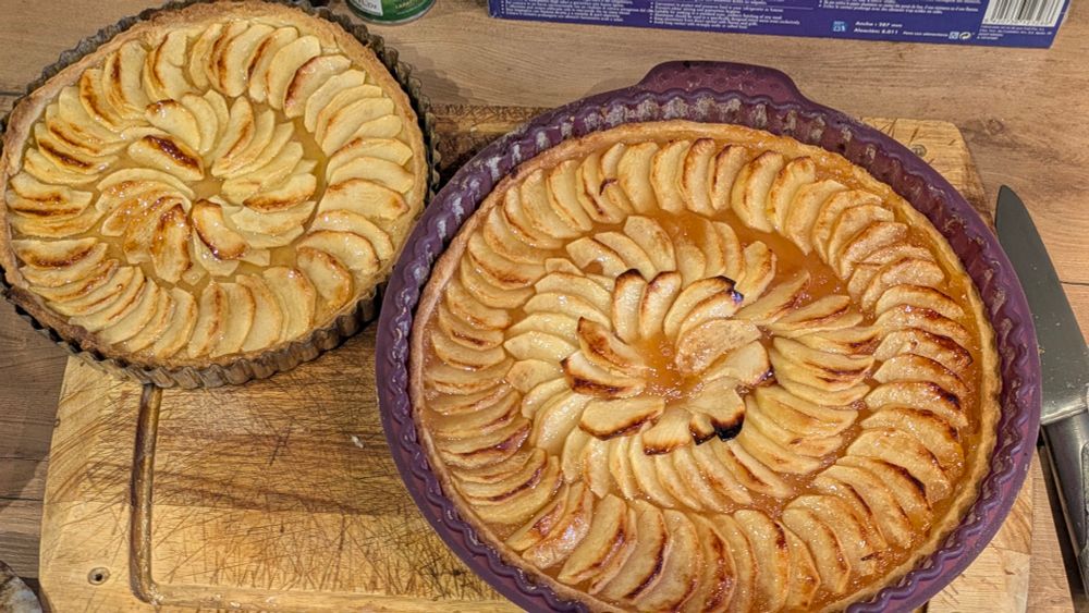 Une paire de tartes aux pommes posées sur une planche en bois. Celle de gauche n'a pas de fond de compote et est dans un moule en metal de 20cm, celle de droite dans un moule en silicone de 26cm a un fond de compote