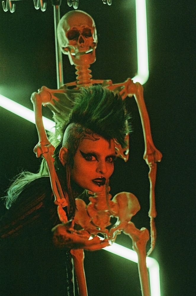 a person with a green mohawk and dramatic makeup makes a scared face whilst being embraced by a skeleton on a stand, lit in red with a greenish backlight