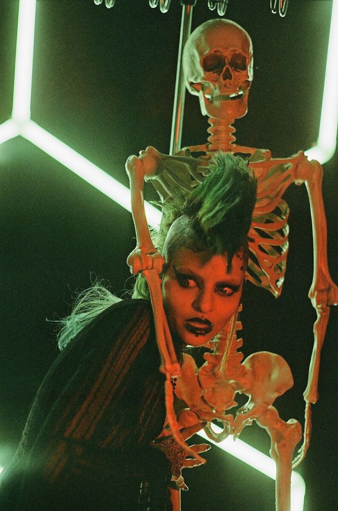 a person with a green mohawk and dramatic makeup makes a scared face whilst being embraced by a skeleton on a stand, lit in red with a greenish backlight