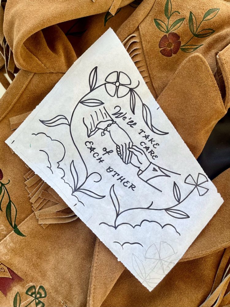 A close cropped photo of a warm-toned suede jacket with fringe, hinted with hand painted flowers, and a piece of paper laid on top. The paper has a drawing of two hands holding each other and the words “we’ll take care of each other” framed by clover and a flower growing on vines. It’s implied that this drawing will be painted onto the jacket below.