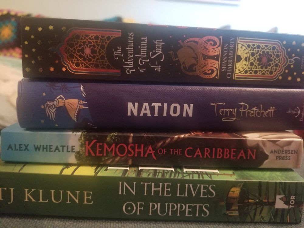 Stack of books. The Adventures of Amina al-Arian by Shannon Chakraborry, Kemosha of the Caribbean by Alex Wheatle, Nation by Terry Pratchett and In the lives of Puppets by T J Klune.