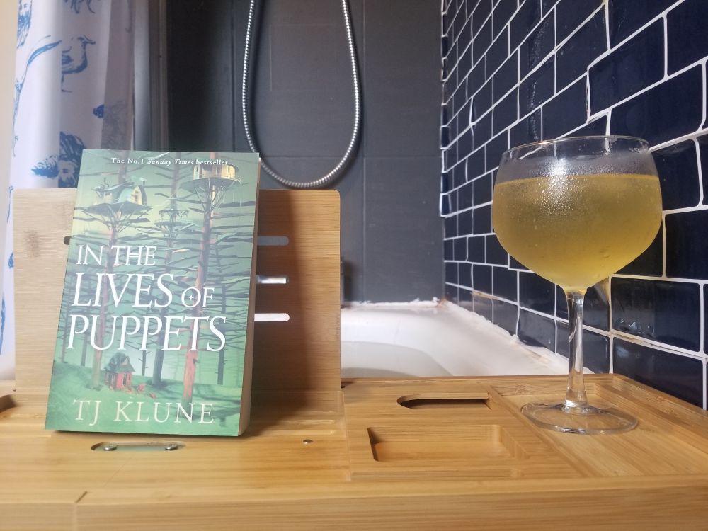 A bamboo bath tray over a white bathtub. One wall is dark blue tiles and the other grey. A rum cocktail and a copy of In the lives of Puppets by TJ Klune sit on it. You can just see my blue on white Lucia's sketchbook shower curtain. 