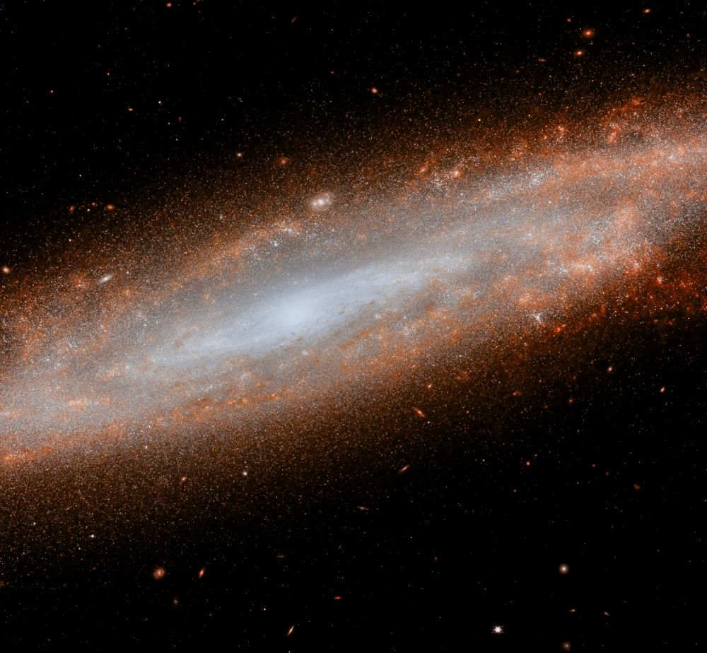 A spiral galaxy, NGC-4666, seen in infrared light. The centre is bright and mostly white. The arms are seen in red and orange colours. The galaxy spreads from the left side of the image onto the right. Several background galaxies can be spotted on the upper and bottom parts of the image (even through the arms of NGC-4666!).