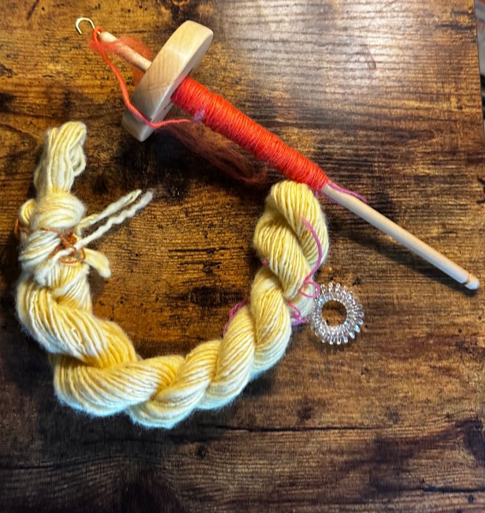 My very first handspun skein next to my second on a wooden top whorl spindle that’s in progress. The finished skein is yellow Corriedale woolen yarn. The yarn one on the wooden spindle is deep coral Corriedale wool. 