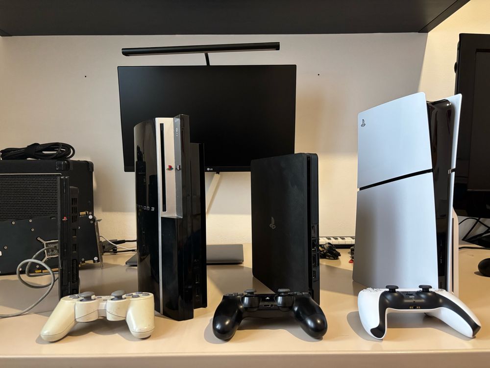 A Playstation 2 slim, a Playstation 3 fat, a Playstation 4 slim and a Playstion 5 slim, sitting on a desk in front of their respective controller.