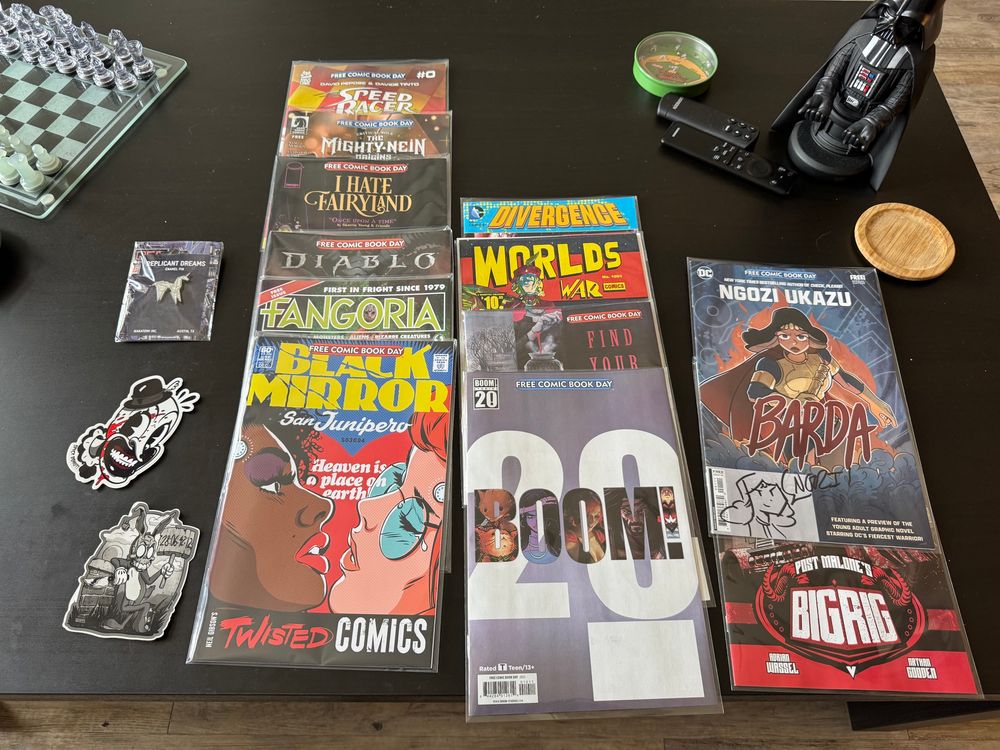 Overhead photo of a black coffee table showcasing my Free Comic Book Day haul, including, from top-left to bottom-right: an enamel pin of the origami unicorn from Blade Runner by Tim Doyle, a sticker of Art the Clown by Spicy Donut, a sticker of Frank in his Bunny costume by Spicy Donut, Speed Racer issue #0, The Mighty Nein Origins #0, I Hate Fairyland “Once Upon a Time”, Diablo: Dawn of Hatred, signed copy of Fangoria Free Comic Book Day 2025 special, Black Mirror: San Junipero, Divergence issue #0, Worlds War #1001, Find your Dark New Feat, Boom! Studios 20th anniversary collection, signed copy of Barda Free Comic Book Day edition, and Post Malone’s Big Rig issue #0